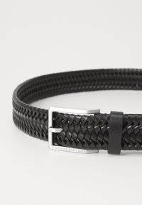 Musta punottu nahkavyö hopeisella metallisoljella. Varustettu sileällä pinnalla ja solkeen on kaiverrettu logo.