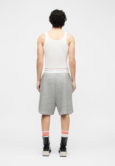 Άνδρας με σγουρά μαλλιά, ντυμένος με λευκή φανέλα, γκρίζα μακριά σορτς, λευκές κάλτσες με πορτοκαλί ρίγες και μαύρα μποτάκια, στέκεται με την πλάτη στραμμένη.