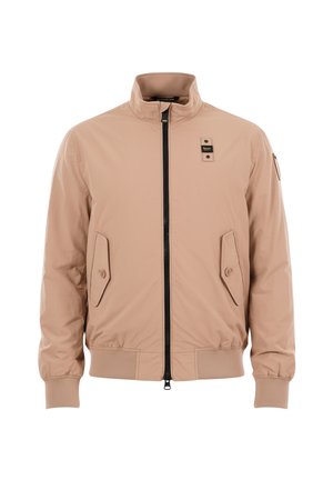 Giacca beige con zip, collo alla coreana, due tasche frontali con patta e bottone, polsini e orlo a costine, e una piccola toppa sul petto e sulla manica.