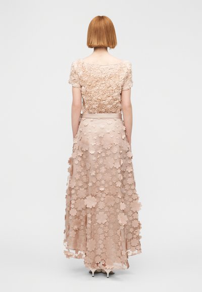 Φόρεμα Maxi σε απαλό μπεζ χρώμα με υφασμάτινο μπούστο με λουλουδένιο ανάγλυφο και εφαρμογές 3D λουλουδιών, καθώς και φούστα με στρώσεις και διαφανές επικάλυμμα.
