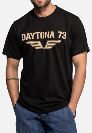Homme portant un t-shirt noir avec « DAYTONA 73 » et un logo d'aile stylisé en beige, associé à un jean bleu.