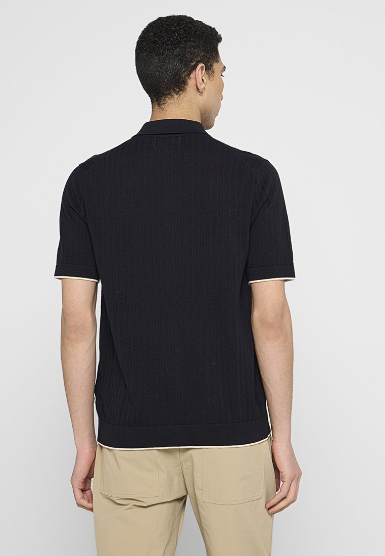 Koszulka polo z krótkim rękawem w kolorze czarnym, z teksturą ryżową, kremowymi akcentami na mankietach i kołnierzyku oraz luźnym krojem. Pokaź widok z tyłu.
