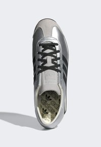 Silver athletic shoe with black laces, suede and synthetic materials, three black stripes, padded tongue, and logo-patterned insole.