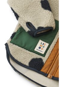 Fleece bunda s krémovým tělem, tmavými skvrnami, zelenými a hnědými panely, se zipovým uzávěrem a nášivkou. Měkká textura, kulatý tvar.