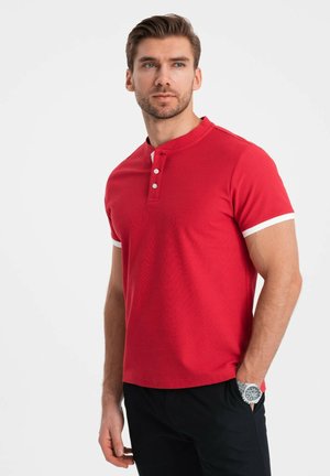 Rød polo-shirt med knaplukning, hvid kant på ærmerne, korte ærmer og et struktureret stof. Bæres med sorte bukser.
