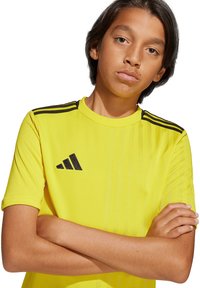 Gele sportshirt met zwarte strepen op de schouders. Bevat het Adidas-logo in het zwart. Gemaakt van lichtgewicht, ademend materiaal.