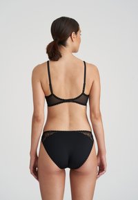 Svart underklädesset med en genomskinlig mesh-bh med vågiga kanter och matchande trosor med spetsdetaljer på sidorna. Visat bakifrån.