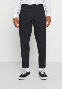 Muži v tmavě navy chinos kalhotách s vyhrnutými kotníky, bílých ponožkách, černých teniskách s bílými tkaničkami a bílém tričku s dlouhým rukávem.