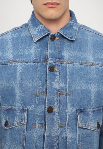 Denim jacket in light blue with faded script patterns. Features a traditional collar, six buttons, and two chest pockets with flap closures.