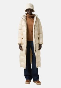 Een persoon met een donkere huid die een lange crèmekleurige pufferjas, een bruine turtleneck trui, breedbenige jeans, tan kleurige chunky schoenen en een crèmekleurige gewatteerde bucket hat draagt.