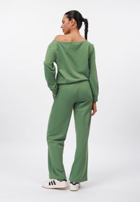 Completo verde a due pezzi realizzato in un tessuto morbido; top off-the-shoulder e pantaloni a vestibilità ampia con tasche; indossato con scarpe bianche con strisce nere.