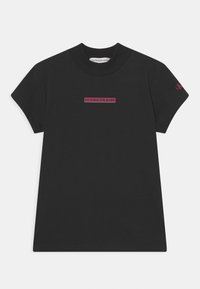 Camiseta de algodón negra de manga corta. Presenta un logo rectangular en texto rojo "CALVIN KLEIN JEANS" en la parte frontal y un pequeño logo en la manga.