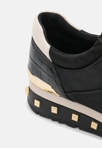 Talons de baskets noirs et beiges avec des clous dorés sur la semelle, un accent doré et un matériau noir texturé sur la partie supérieure.