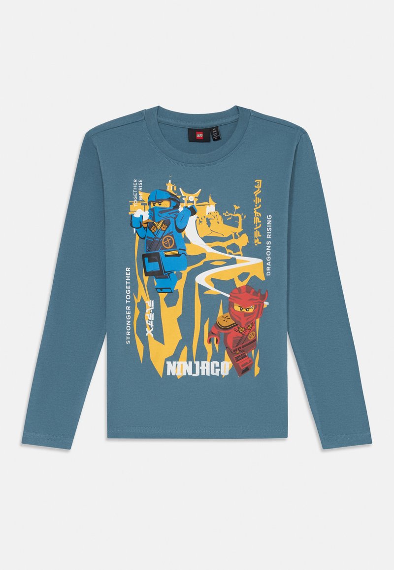 Langermet blå bomull T-skjorte med gule og røde ninja-grafikker og "Ninjago"-tekst, satt mot et stilisert bakgrunn.