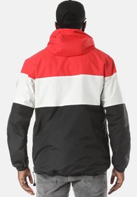 Uomo che indossa una giacca a vento tricolore rossa, bianca e nera, con un berretto nero, in piedi di spalle con le braccia rilassate lungo i fianchi.