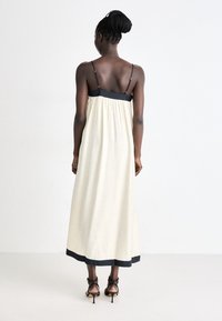 Lang, cremefarbendes Kleid mit schwarzem Taillenbund und Saum, ausgestattet mit verstellbaren Trägern. Glatte Textur, fließendes Design. Es werden schwarze Stilettos getragen.