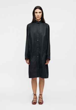 Γυναίκα με ίσια σκουρόχρωμα μαλλιά φορεί ένα μαύρο παλτό μέχρι τα γόνατα με κουμπιά και καφέ δερμάτινα δετά παπούτσια με χαμηλή σόλα, στέκεται μπροστά σε ένα απλό λευκό φόντο.