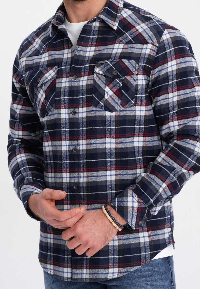Homme portant une chemise à carreaux bleu marine, blanche et rouge boutonnée avec deux poches poitrine, un jean bleu et des bracelets de perles au poignet gauche.