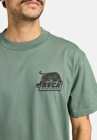 Groene katoen t-shirt met een zwart leeuw-graphic en de tekst "RVCA NATURE VS INDUSTRY" op de linkerkant van de borst. Korte mouwen, ronde hals.