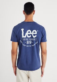 Granatowa koszulka z krótkim rękawem z bawełny, z dużym białym logo na plecach z napisem "Lee Salina, Kansas 89 Stworzona z myślą o celu."