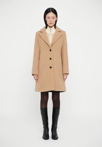 Abrigo de lana beige con cuello de muesca, tres botones negros y forma ajustada, usado sobre un suéter crema, combinado con botas negras hasta la rodilla.