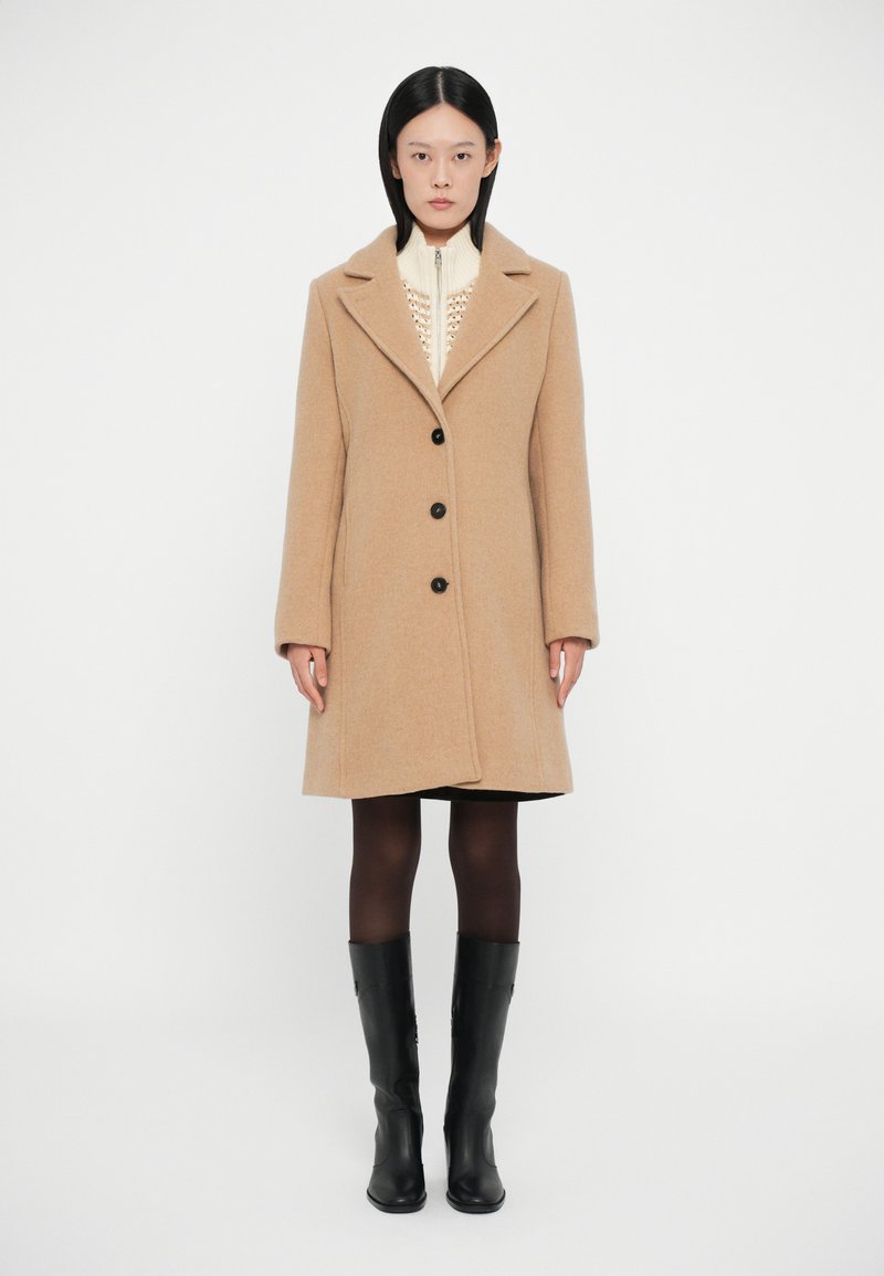 Abrigo de lana beige con cuello de muesca, tres botones negros y forma ajustada, usado sobre un suéter crema, combinado con botas negras hasta la rodilla.
