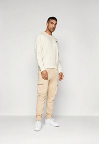 Beige Pullover mit Rundhalsausschnitt und Logo, kombiniert mit passenden Cargo-Hosen mit Seitentaschen, vervollständigt durch weiße Turnschuhe.