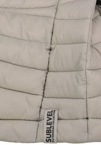 Helles graues Puffergewebe mit einem gesteppten Muster. Verfügt über ein SUBLEVEL-Tag. Das Material wirkt glatt mit subtilen vertikalen Streifen.