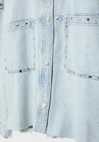 Section de veste en jean bleu clair avec deux poches avant ornées de rivets argentés et des boutons-pression métalliques visibles en plein centre.