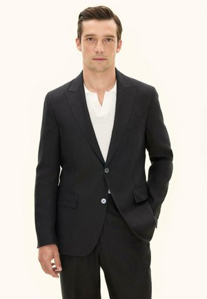 Mand iført en sort dragt over en hvid tekstureret skjorte, står med den ene hånd i lommen, neutralt baggrund.