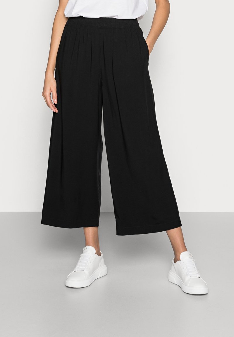Personne portant un pantalon court noir à jambes larges, des baskets blanches et un haut blanc avec une main dans la poche.