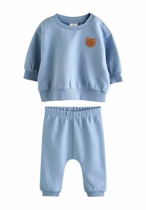 Jasnoniebieski strój dziecięcy z bluzką na długi rękaw ozdobioną małą brązową łatką w kształcie misia oraz pasujące spodnie z elastyczną talią.