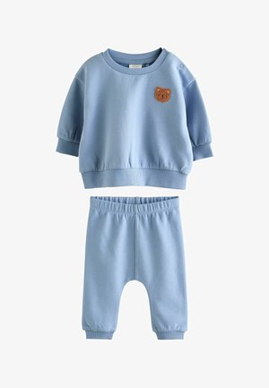 Svetlo modra otroška obleka z dolgimi rokavi, na vrhu je majhen rjav krzneni medvedek, hlače pa imajo elastičen pas.