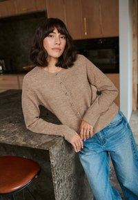 Cardigan en tricot marron avec des boutons argentés, porté sur un jean large bleu clair, devant un plan de travail en pierre texturée dans une cuisine moderne.