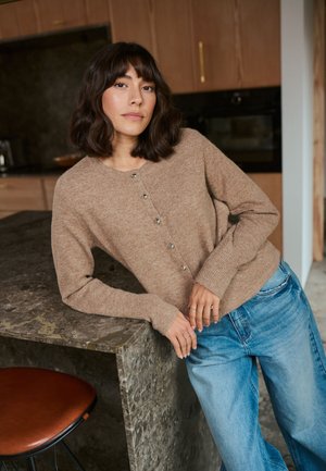 Brauner Strickcardigan mit silbernen Knöpfen, getragen über hellblauen weiten Jeans, vor einer strukturierten Steinarbeitsplatte in einer modernen Küche.