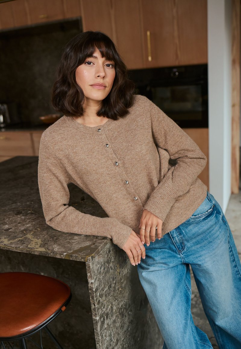 Cardigan en tricot marron avec des boutons argentés, porté sur un jean large bleu clair, devant un plan de travail en pierre texturée dans une cuisine moderne.