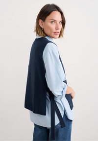 Donkerblauwe gebreide vest met open zijkanten, geribbelde zoom en strikjes bij de taille, gedragen over een lichtblauwe gestreepte blouse en denim jeans.