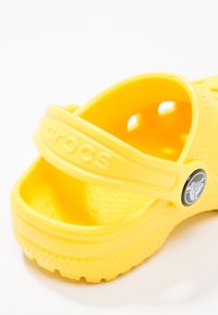 Sandale en caoutchouc jaune avec sangle ajustable, perforations pour la respirabilité, semelle texturée et accent de logo circulaire.