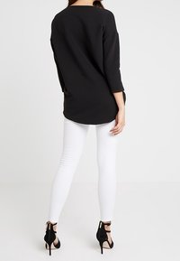 Musta pitkähiha toppi, jossa on teksturoitu pinta, yhdistettynä istuviin valkoisiin housuihin ja mustiin korkokenkiin. Takaviesti, jossa on pyöreä helma.