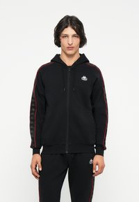 Sudadera con cremallera negra, franjas laterales rojas, bolsillos tipo canguro y capucha con cordón. Presenta un logo en el pecho y detalles estampados en las mangas.