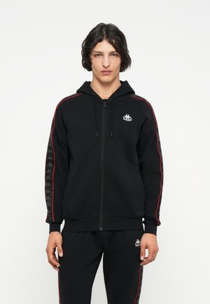 Sudadera con cremallera negra, franjas laterales rojas, bolsillos tipo canguro y capucha con cordón. Presenta un logo en el pecho y detalles estampados en las mangas.