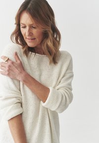 Hvid strikket sweater med en løs pasform og omviklede ærmer, der har et struktureret mønster. Modellen bærer en guldring på den ene hånd.