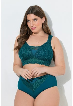 Donna curvy che indossa un reggiseno in pizzo turchese e slip a vita alta abbinati, in piedi su uno sfondo bianco, con le mani unite in vita.