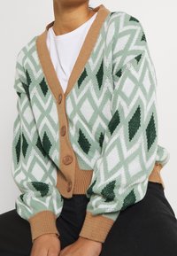 Sweter kardigan w zielono-białe wzory z beżowym przodem zapinanym na guziki, zdobiony geometrycznymi diamentowymi kształtami i wykończony prążkowanymi mankietami.