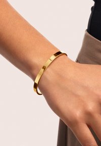 Bracciale in metallo dorato con una finitura liscia e lucida. Presenta un design minimalista e aperto che avvolge il polso.