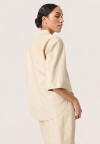 Beige læderbluse med korte ærmer, en afslappet pasform og buet kant. Har en glat overflade og minimale syninger.
