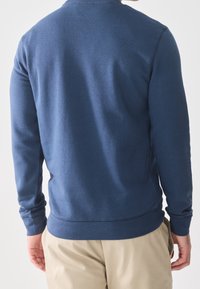 Mand iført en mørkeblå langærmet sweater og beige bukser, set bagfra mod en ensformet hvid baggrund.