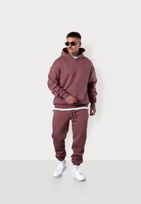 Bordeauxrotes Baumwoll-Hoodie mit einer Fronttasche und einer passenden Jogginghose, kombiniert mit weißen Sneakern und runden Sonnenbrillen.