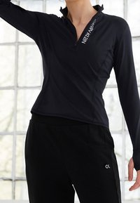 Žena v černém svetru na zip s dlouhým rukávem a nápisem "CALVIN KLEIN" na límci, a černých kalhotách s malým logem "ck" na stehně.