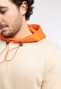 Sudadera beige con cordón y cuello naranja; presenta el logotipo blanco "FORSBERG". Tejido suave con un corte relajado y detalles de costura visibles.